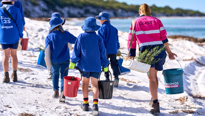 Children in school uniform carry buckets of seedlings along a beach, helped by a Shire officer in hi-vis work gear, carrying more seedlings