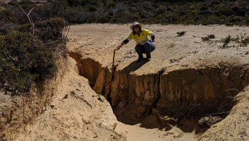 A member of the Shire Environmental team crouches at the top of a section of severely eroded 4WD track, demonstrating how deep it is with a long handled shovel. She is holding the very top of the shovel just above ground height and the bottom of the shovel rests on the bottom of the erosion.